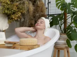 A woman relaxing in a bathtub surrounded by plants, with a serene expression on her face, a towel wrapped around her head, and various bathing accessories nearby.