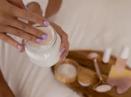 A person using skincare products, seen from above; they scoop cream from a jar with manicured hands, surrounded by a tray of beauty items.