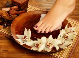 A woman's foot with purple nail polish rests in a wooden bowl surrounded by white orchid flowers, set on a bamboo mat with a dark chocolate brownie and cinnamon sticks nearby.