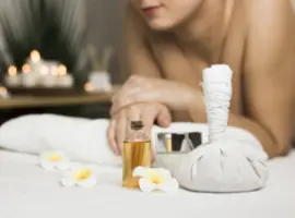 A serene spa setting with a blurred person in the background reaching for a bottle of oil. white towels, a herbal compress, and flowers are crisply focused in the foreground.