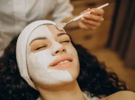 A woman with a headband and closed eyes relaxing while a facial mask is applied to her face with a brush in a spa setting.